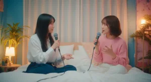 兩位年輕的日本女性坐在房間的床上，正在進行 Podcast 錄製。她們面帶微笑，氣氛輕鬆愉快，畫面具有溫暖的電影感。